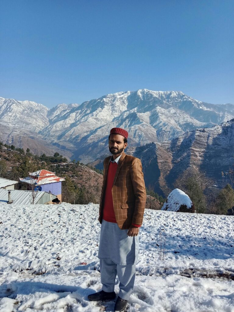 HOME A man stands in traditional attire before sunlit snow-covered mountains.