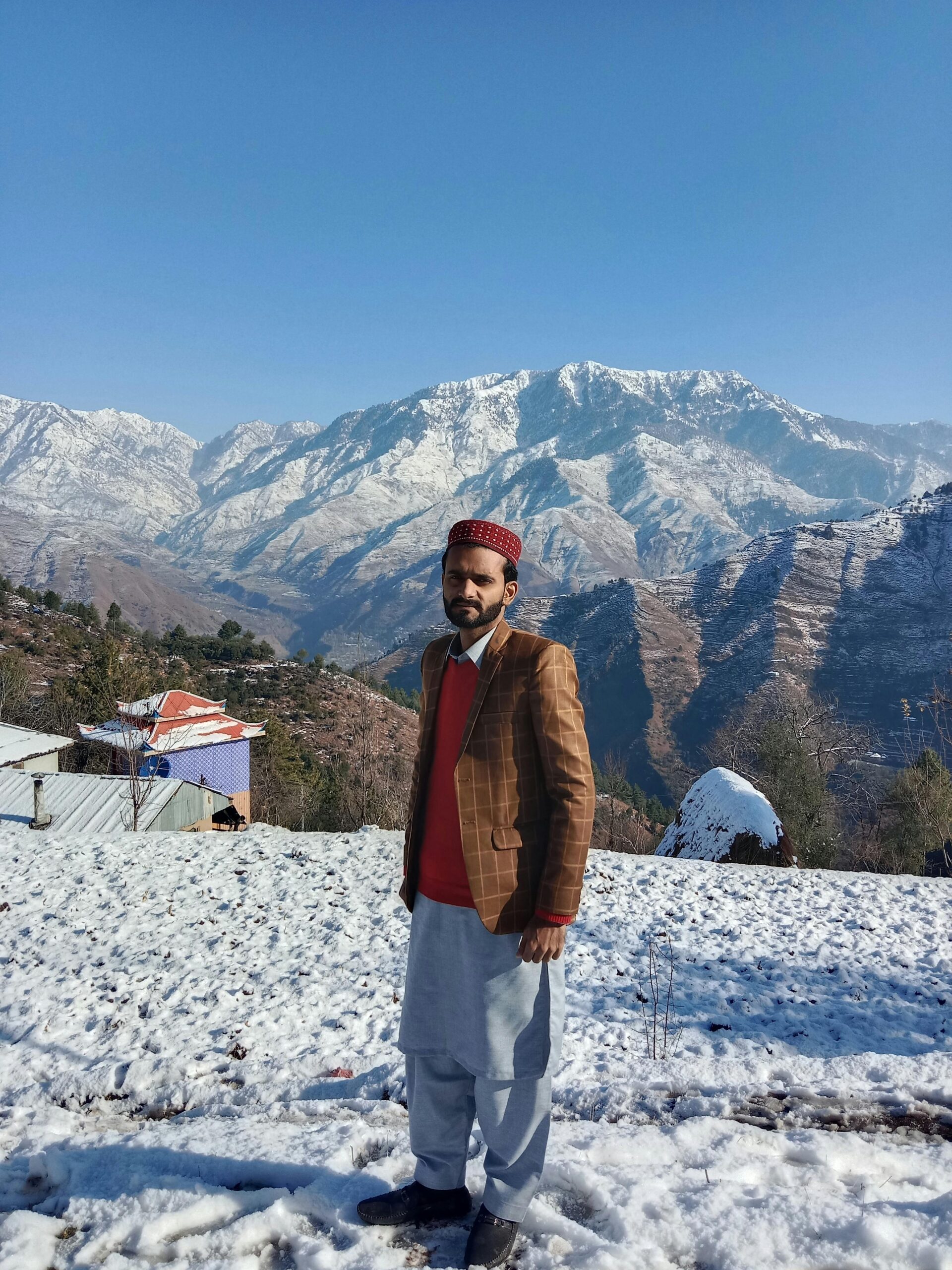 A man stands in traditional attire before sunlit snow-covered mountains.